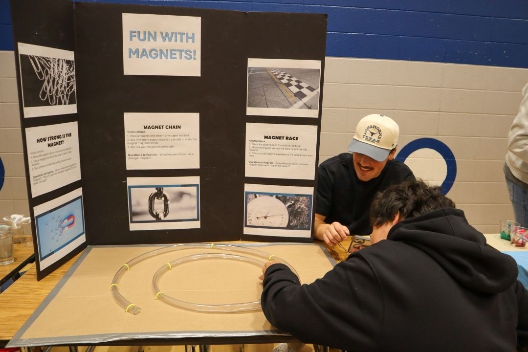 Teacher wearing a hat is showing student how to use magnets using plastic tube on a wooden table with information on tri-fold display board.
